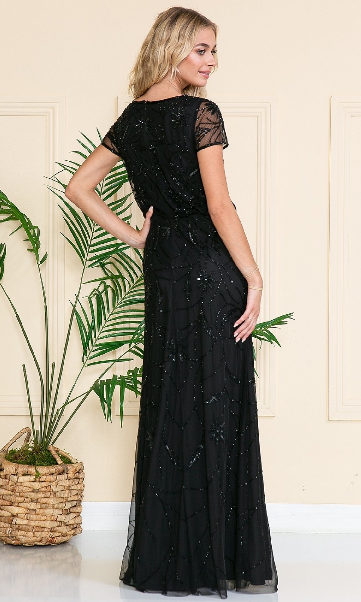 A woman poses indoors near a potted plant, wearing the Amelia Couture IN004 Blouson Long Formal Dress in black, featuring sequin and floral embellishments.